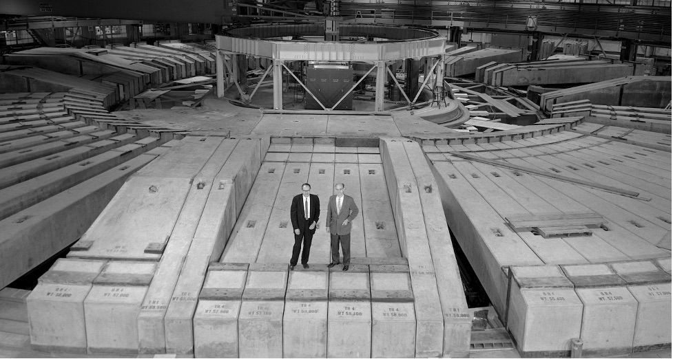 Two men in suits stand amidst a massive structure of concrete blocks inside an industrial facility