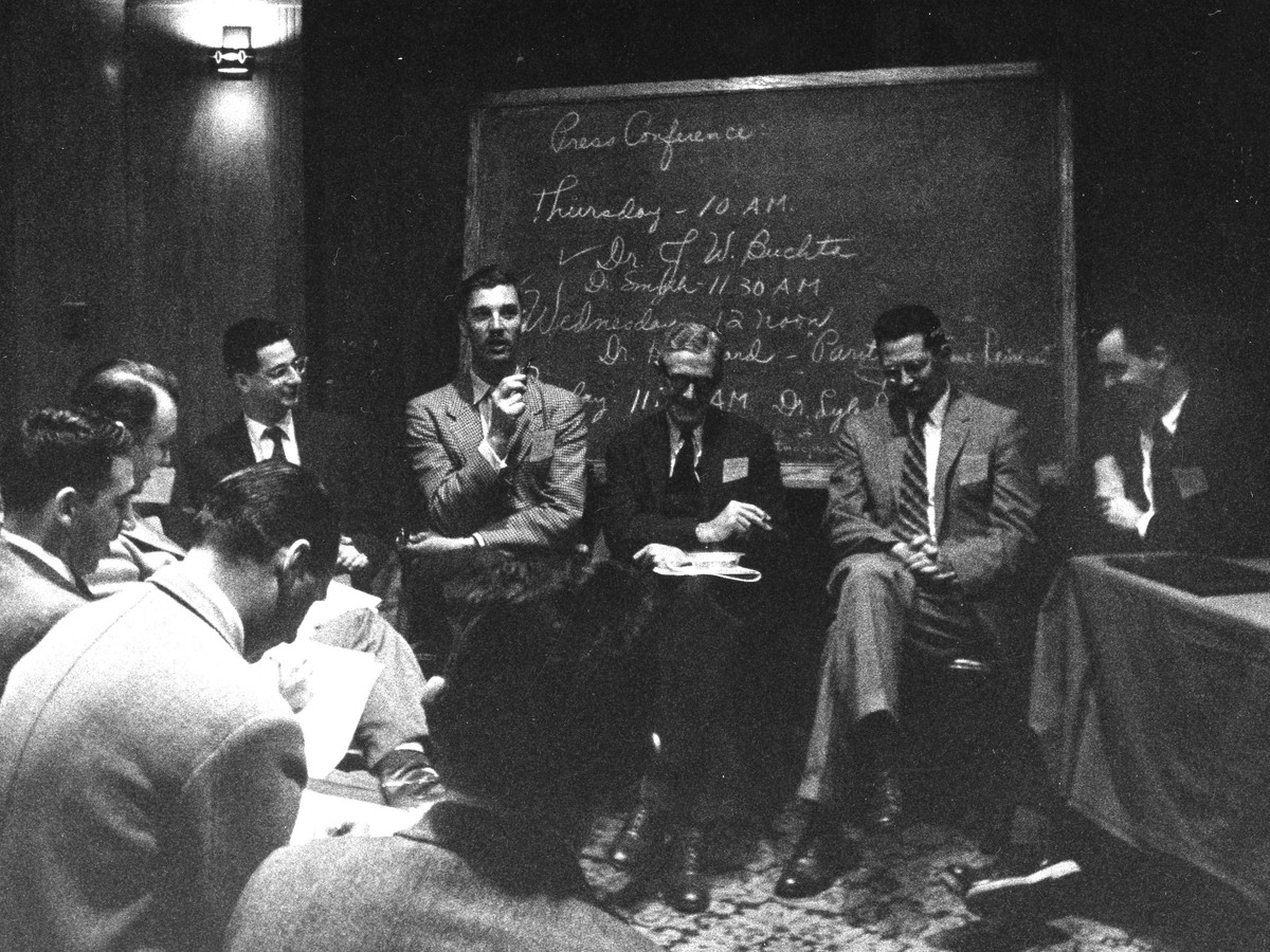 Nine men seated in a circle in front of a blackboard