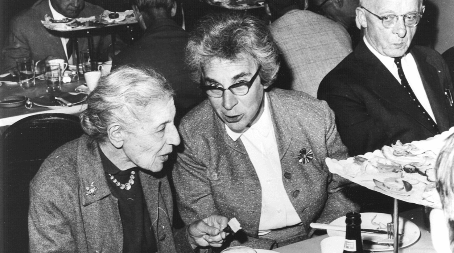 A black-and-white photo depicts two physicists, Lise Meitner and Hilde Levi, who are conversing at a table. Other people are visible in the background.