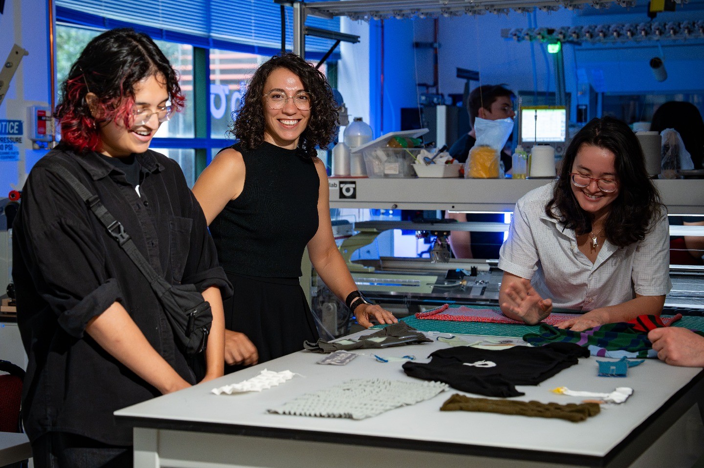 Vanessa Sanchez smiles at the camera while several members of her lab gather around a table with her, looking at fabrics.