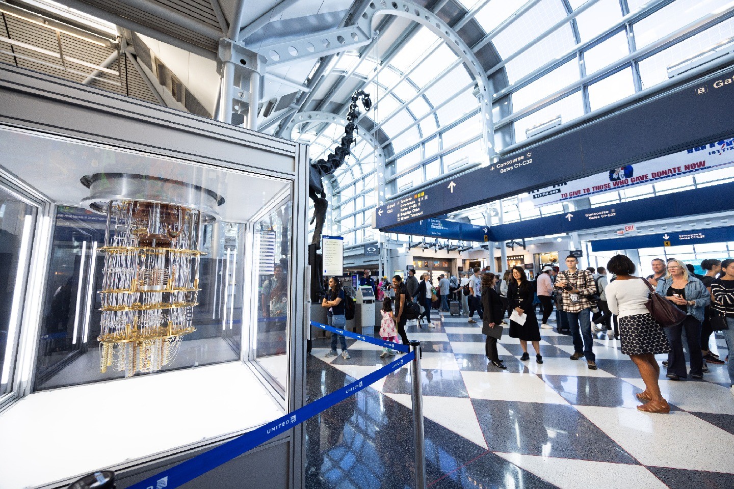 The quantum computer in a glass case is visible, with a large dinosaur fossil in the background, along with airport crowds, signage, and large windows.