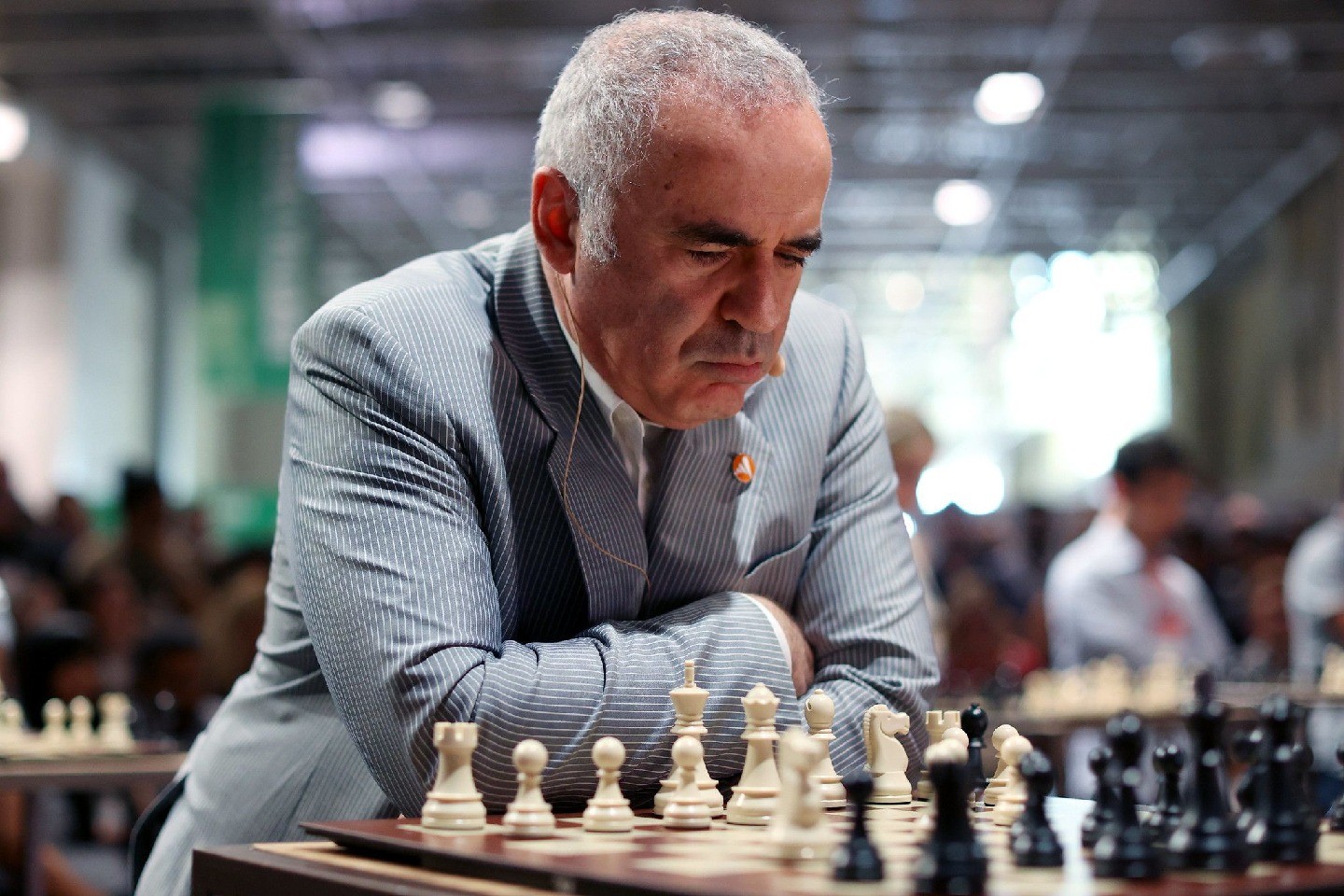 A man sits at a chessboard in a room of others seated at chessboards.