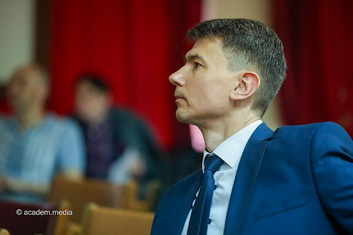 Sergiy Perepelytsya in a blue suit, looking to his right. Behind him are red curtains and two seated figures out of focus.