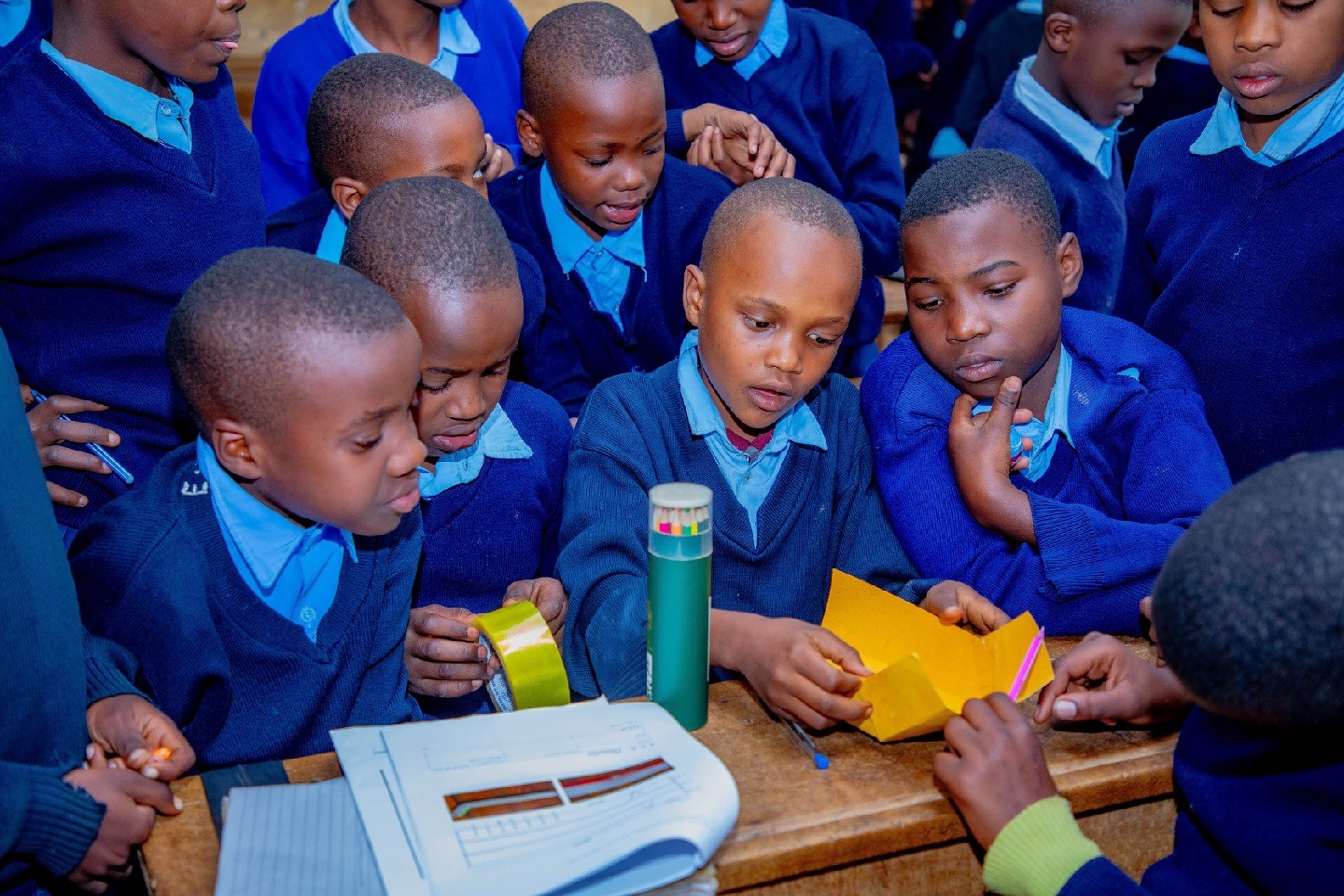 A group of Tanzanian school children wearing blue school uniforms gather around a science experiment on a desk.