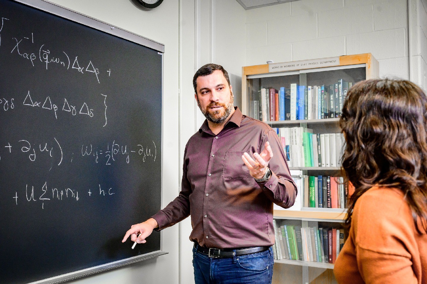 Rafael Fernandes talks to a student next to a blackboard filled with equations.