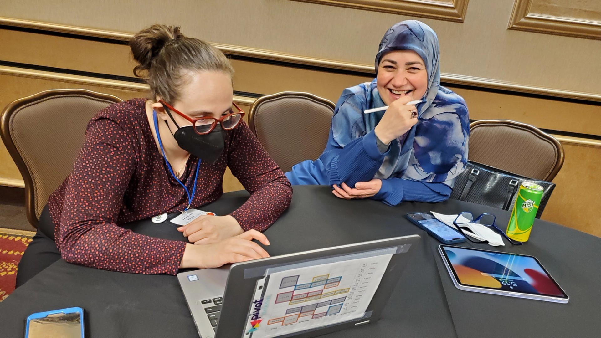 Two women are seated in chairs at a round table. One is focused on a laptop; the other smiles up at the camera.
