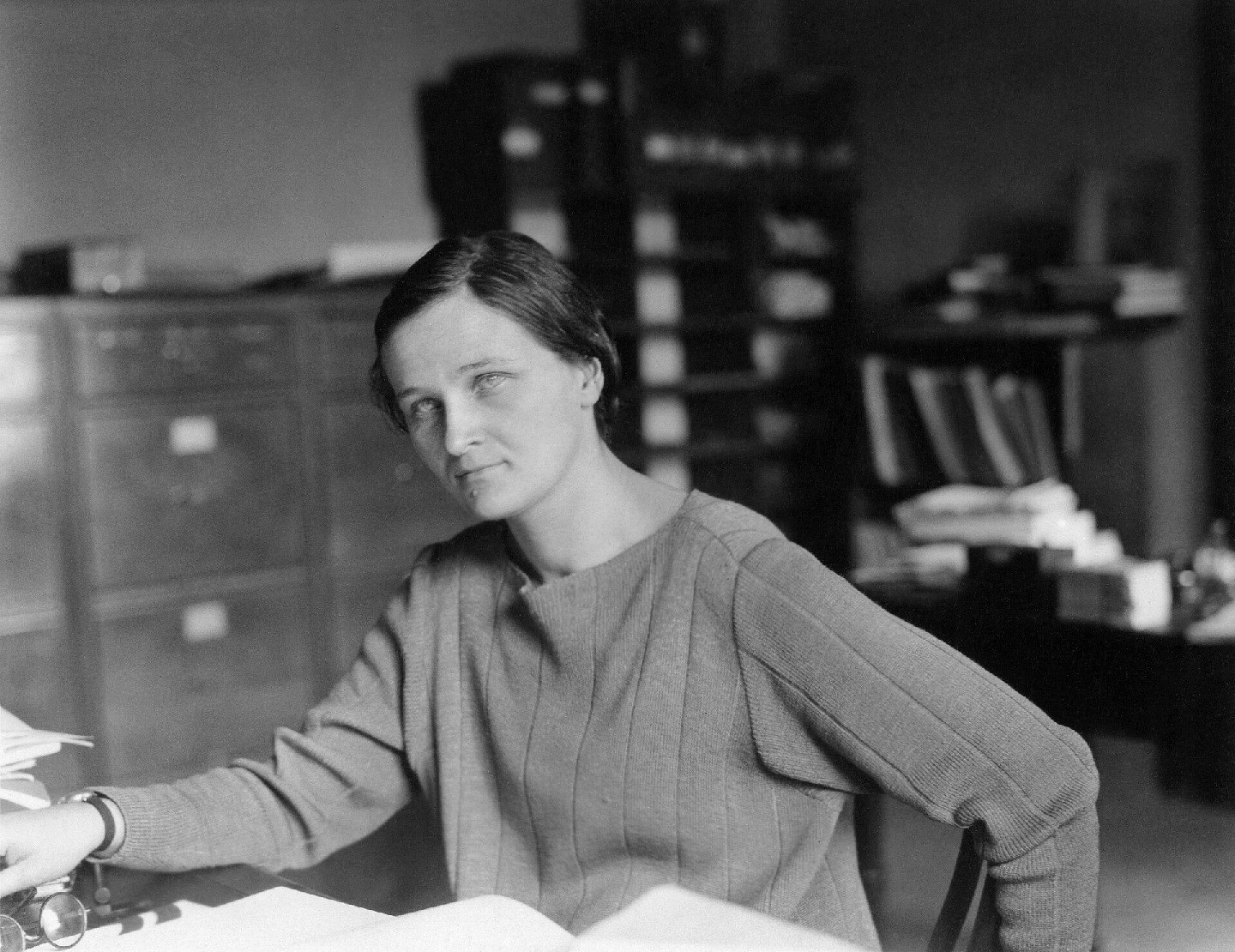 Black-and-white photo of a woman seated at a desk with cabinets behind her.
