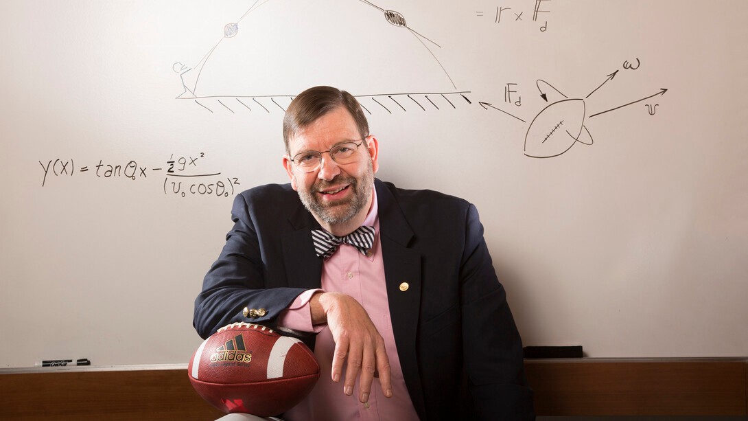 Tim Gay sits with a football in front of a whiteboard with scribbled equations and diagrams related to the physics of football.