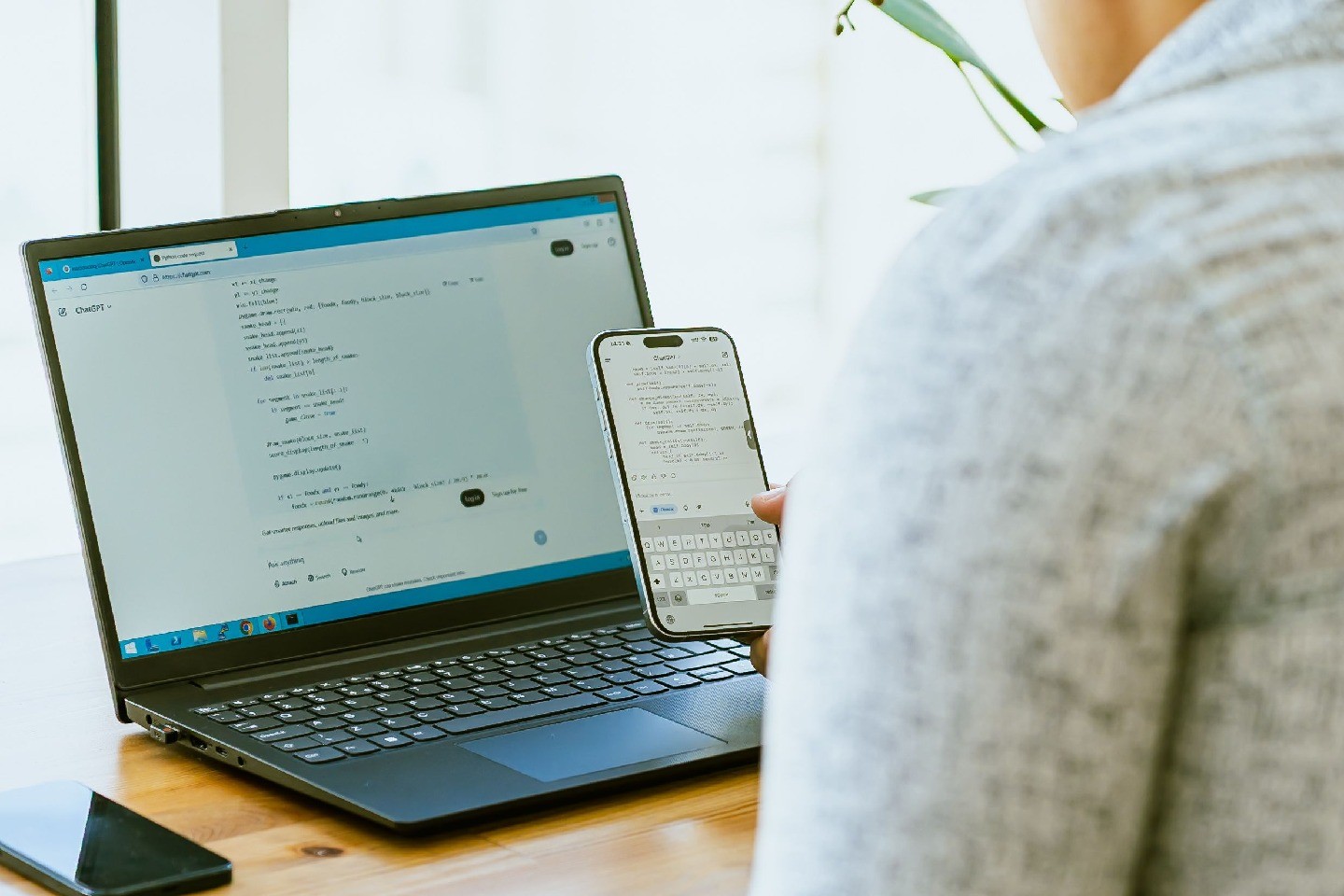 A person holds a phone and looks at a laptop, both with coding visible in AI apps.