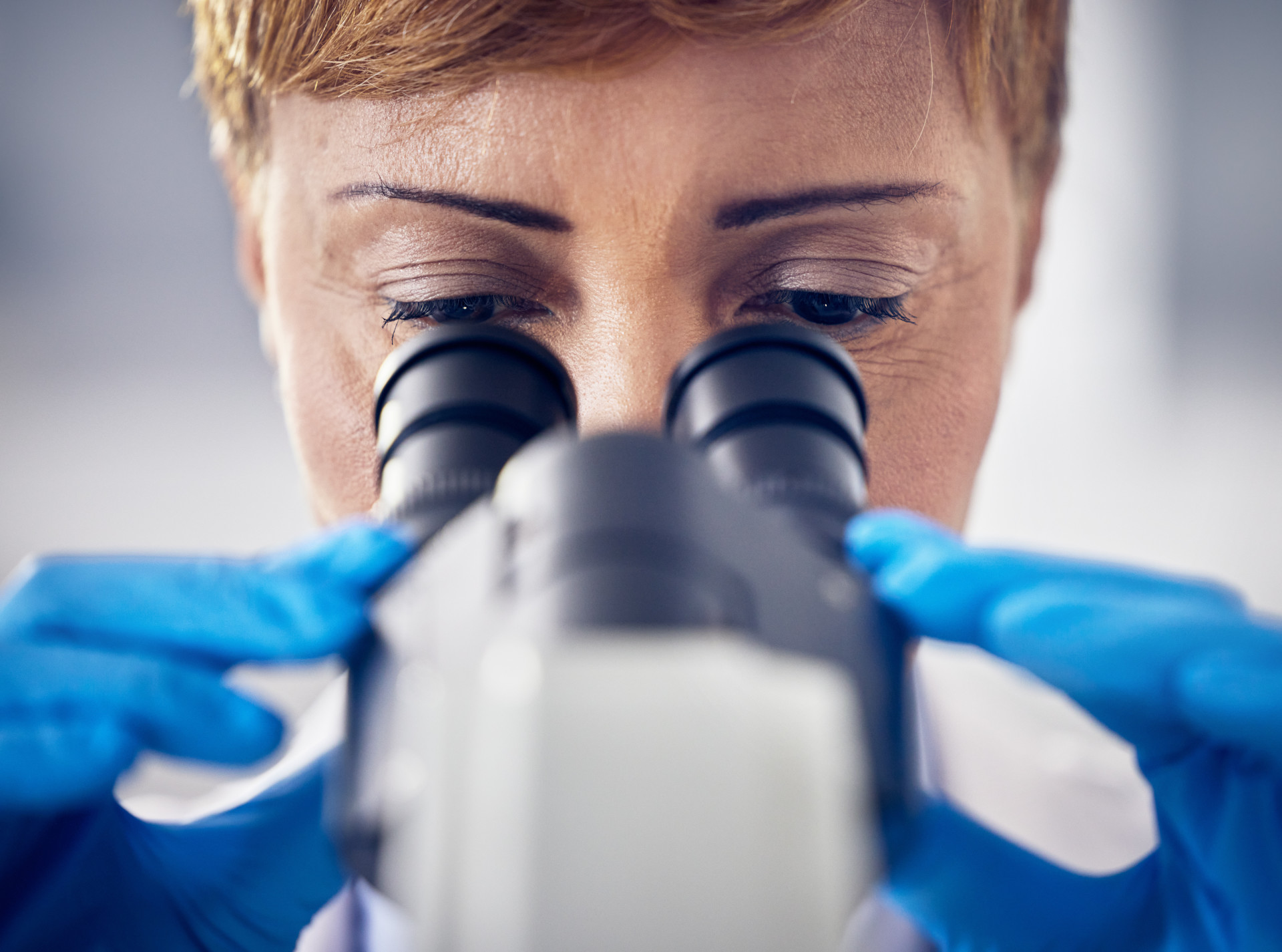 A scientist or healthcare worker wearing blue laboratory gloves and looking through the eyepieces of a microscope. 
