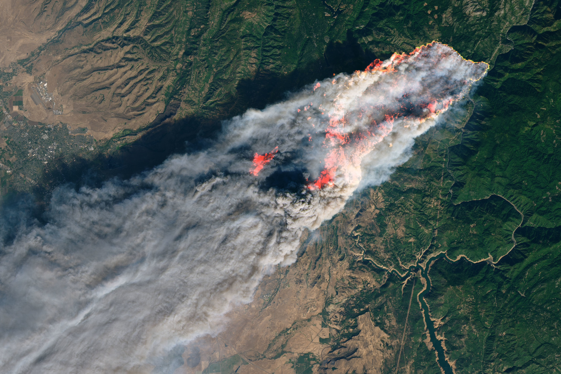 Satellite image of a large fire, showing a massive smoke plume with bright red hot spots marking active fires. Dense white smoke spreads across brown terrain, contrasting with dark green forested areas.