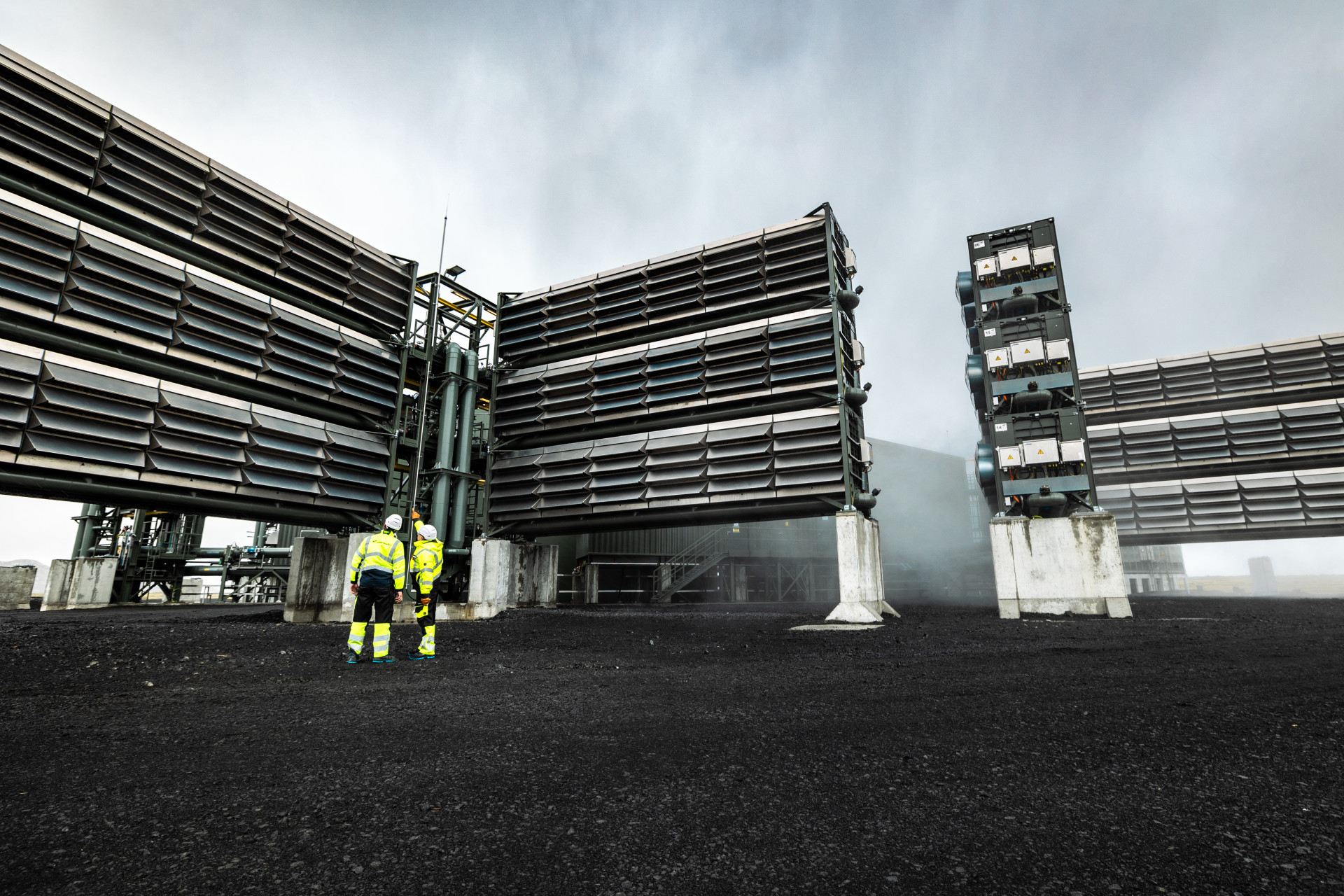 Two workers in yellow safety vests observe large industrial air-capture units with louvered panels mounted on concrete pillars, which contrast starkly with the black ground and overcast sky.