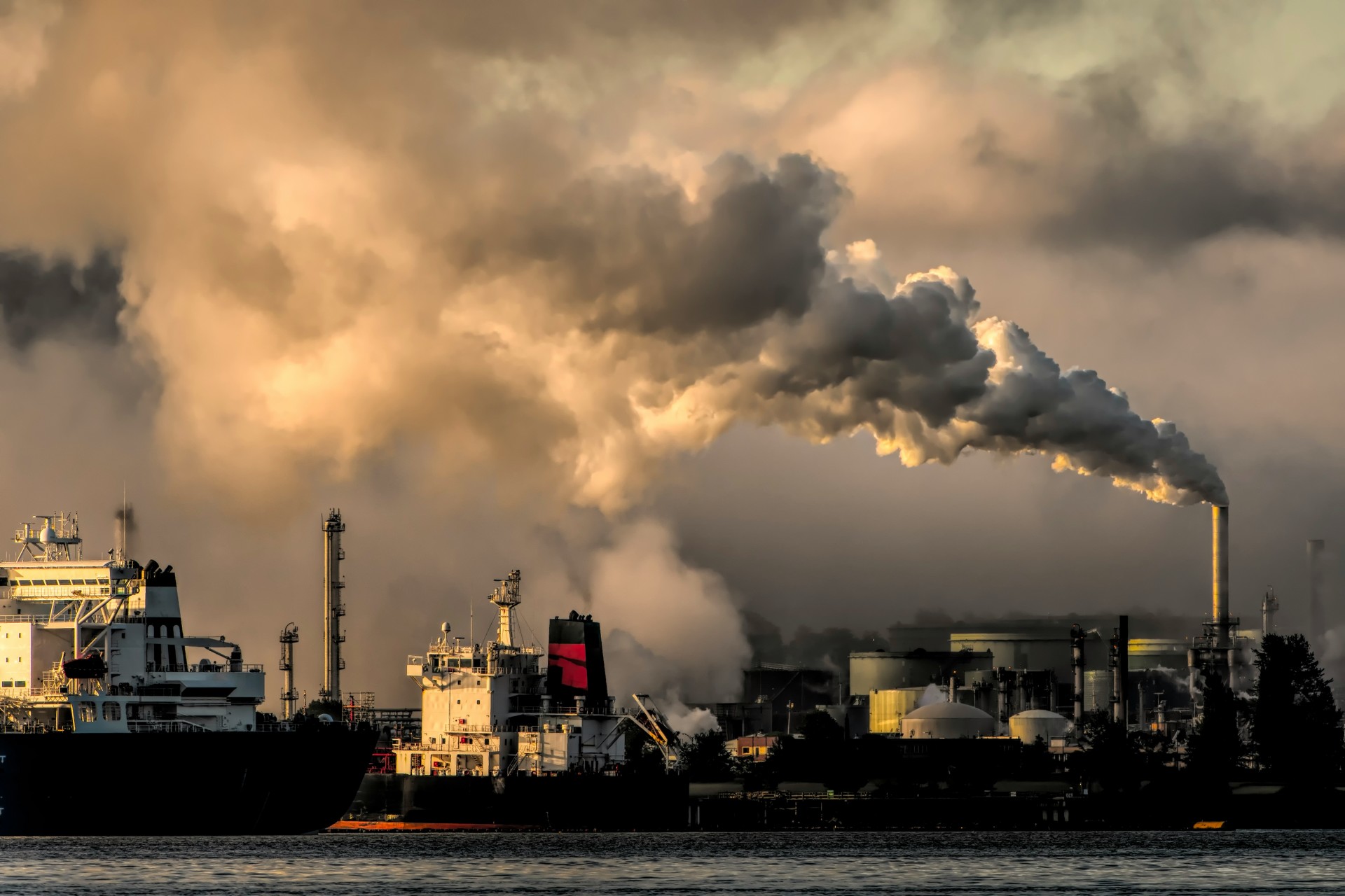 Industrial waterfront shows ships silhouetted against a golden sky, with smoke billowing from a factory smokestack in the background.