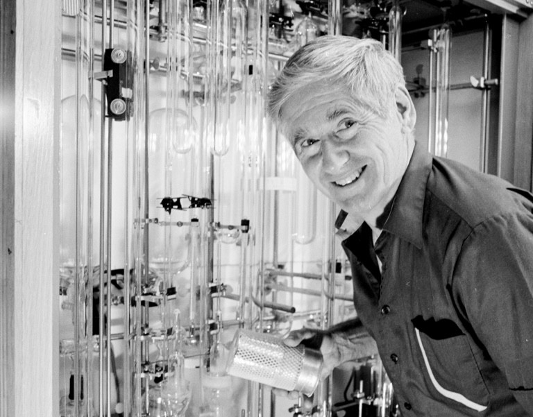 Black and white photo of David Keeling smiling at the camera while working with glass laboratory equipment beside him.