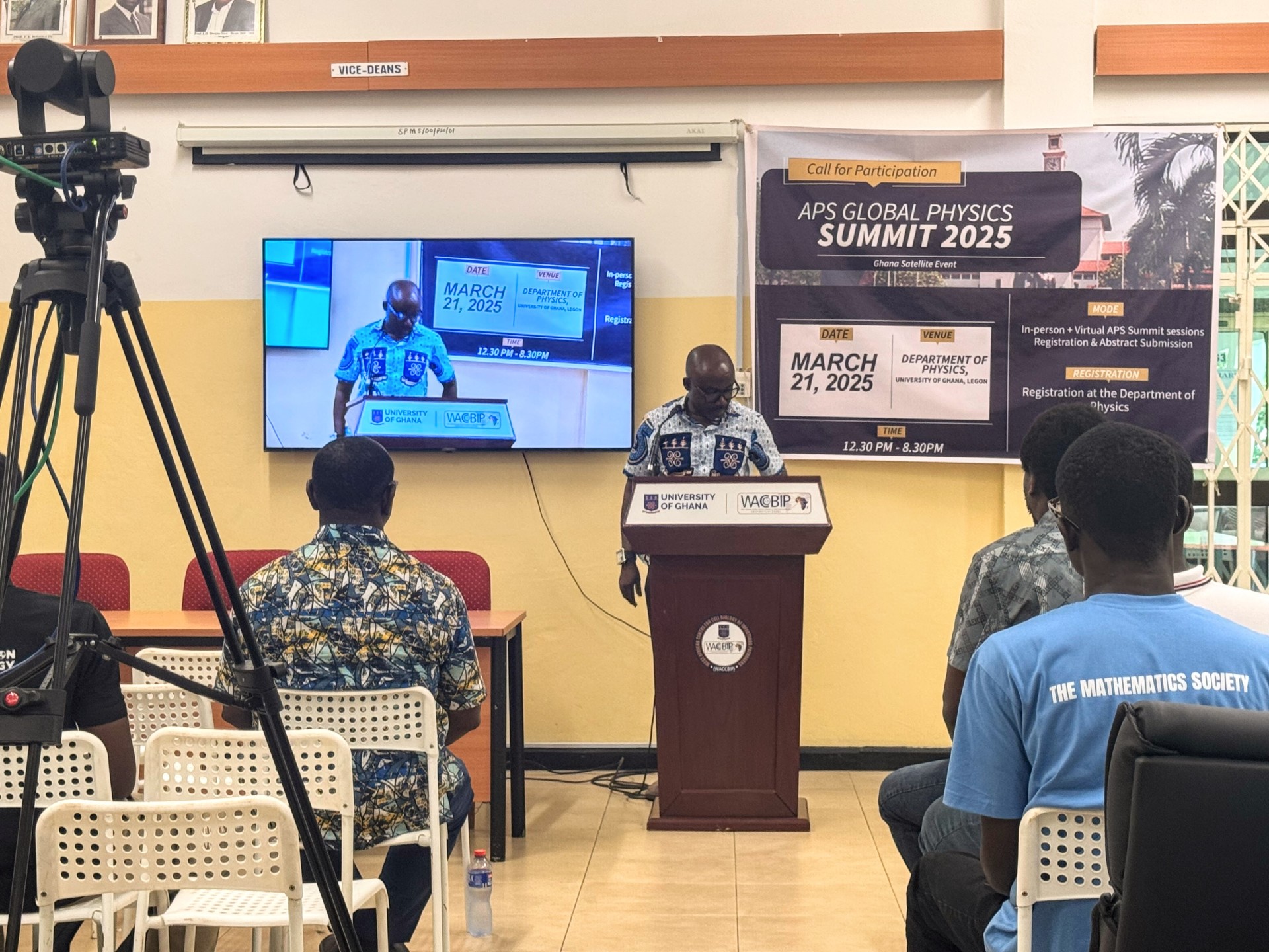 Speaker at a podium presenting, with video feed displayed on screen and attendees watching from chairs