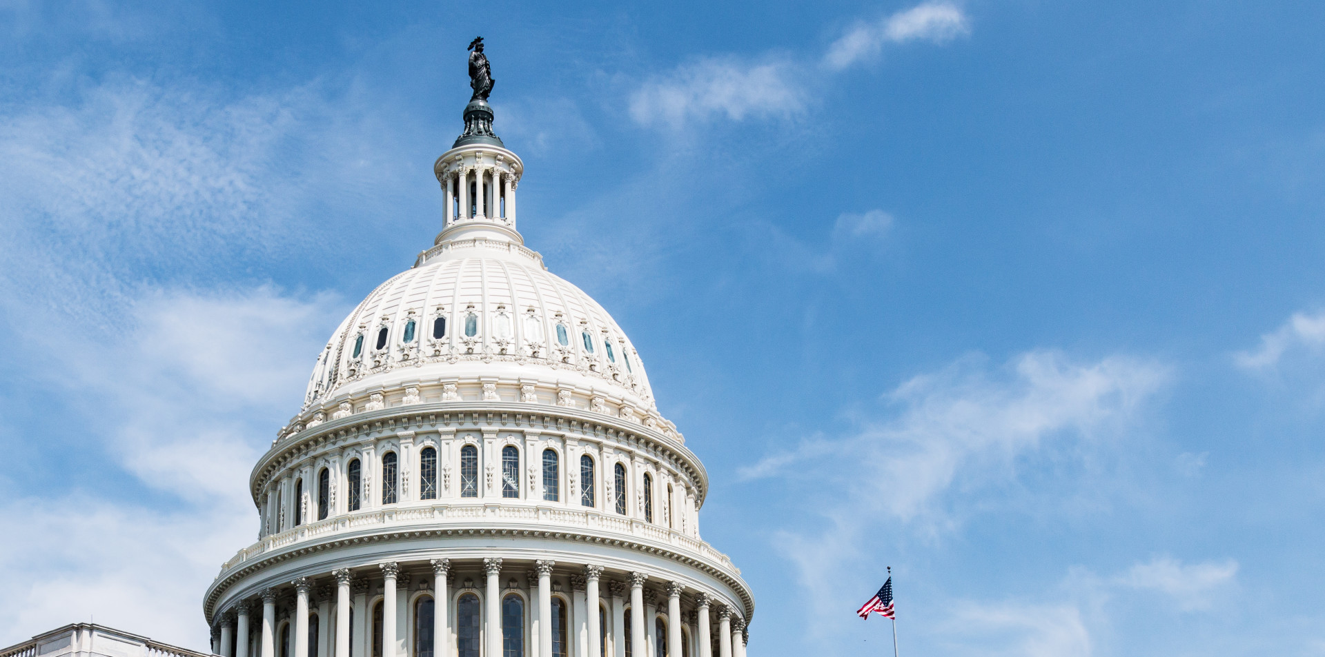 The top of the U.S. Capitol Building with a flag visible and a blue sky above.