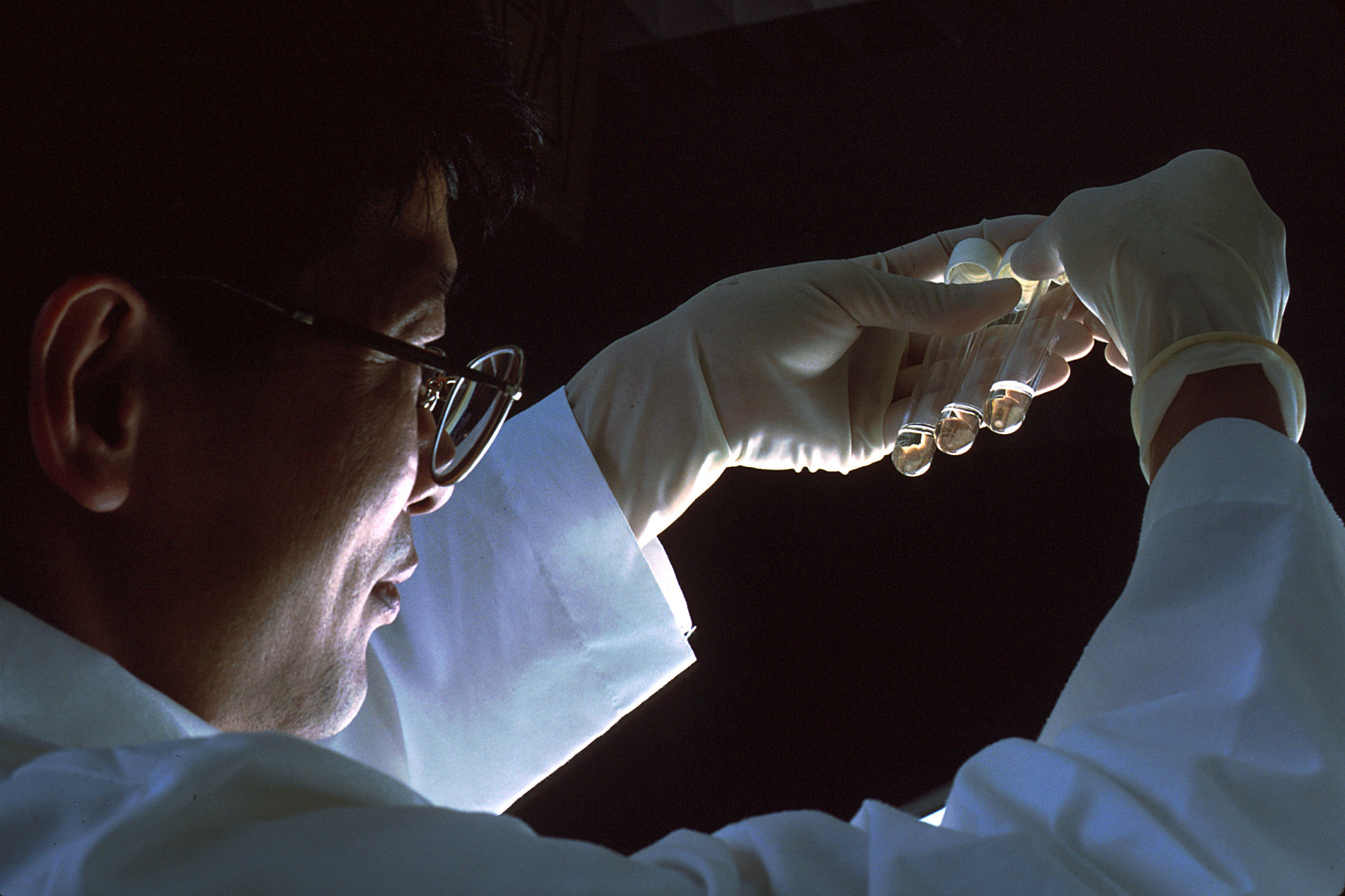 Scientist in glasses and white lab coat examining three small glass test tubes under bright lighting in a darkened laboratory.
