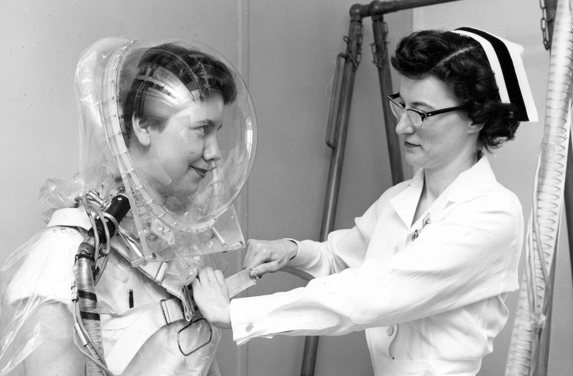 Black and white photo of a nurse in white uniform and cap adjusting equipment on a volunteer wearing a clear plastic dome helmet in a medical laboratory setting.
